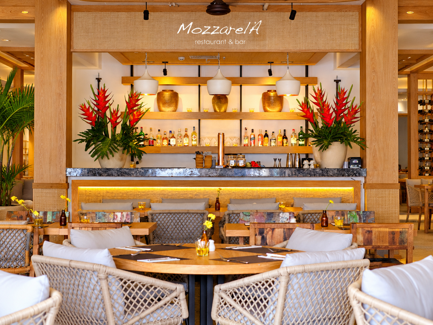 Warm tropical restaurant and bar interior with wooden counter and floral arrangements.